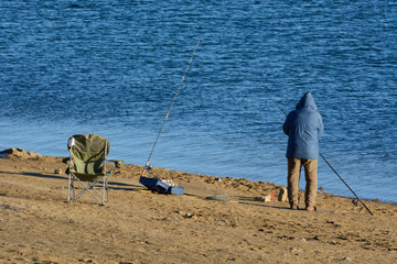 Unrecognizable fisherman fishing alone dressed in winter Unrecognizable fisherman in winter parka with tackle box and chair and several fishing rods fishing alone on lake shore