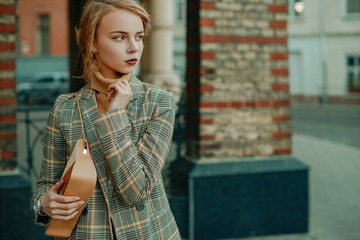 Outdoor portrait of young beautiful fashionable lady wearing stylish checkered blazer, beige cashmere turtleneck, holding small suede bag, posing in street of european city. Copy, empty space for text