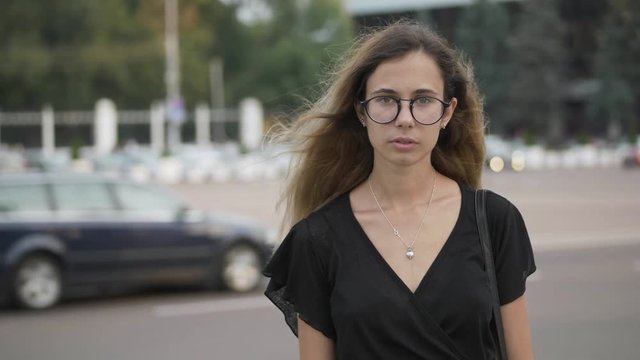 Close up slow motion portrait of young nerdy beautiful caucasian woman in glasses wearing black blouse posing on city street