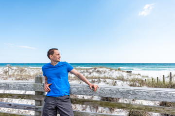 Destin, USA Miramar beach city town village day in Florida panhandle gulf of mexico ocean water, young happy man in blue shirt leaning on wooden fence railing by sand dunes