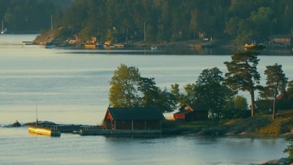Scenic view of a small red house on the rocky shoreline among tall trees. Calm water at sunset. Tranquil, peaceful scene. Slow motion. Camera moving right.