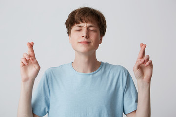 Positive faithful young guy, closing eyes with hope, crossing fingers while making wish, wanting...
