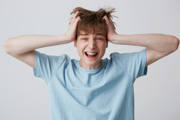 Screaming amazed excited young man clutched at his head, hair tousled, mouth wide opened as shouting loud with braces on teeth wears blue t-shirt, feels happy surprised isolated over white background