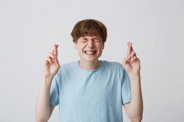 Teenager looks like waiting hoping for something pleasant with screw up eyes, anticipation expression, hands up and fingers twisted, with braces on teeth, wears blue t-shirt over white background