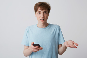 Guy with braces on teeth is looking straight in disbelief, holding a phone in his hand, the second...