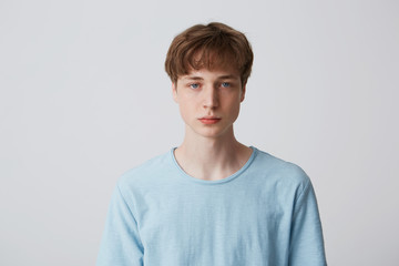 Headshot of a blue-eyed guy without emotions. Emotionless teenager in a blue t-shirt looking to the camera, isolated over white background