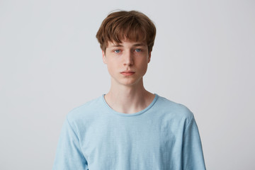 Close up face of a young man without emotions. Beautiful emotionless guy in a blue t-shirt looking to the camera, isolated over white background - obrazy, fototapety, plakaty