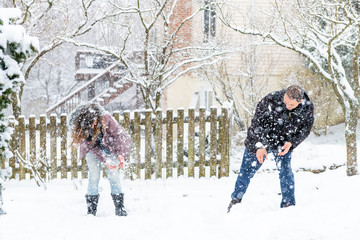 Young woman, man playing, throwing flying snowballs mid-air, air in winter snowstorm, snowing storm at home, house garden, front yard, backyard, trees covered in snow, smiling, happy, laughing