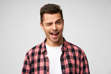 Obraz premium Grinning broadly handsome attractive charming young man, winks with one eye, flirting smile, pleasant appearance, dressed in checkered shirt, standing against white background.