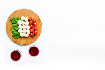 Italian cuisine, food concept. Italian flag made of mozzarella, tomatoes, basil on wooden cutting board near glass of red wine on white background top view space for text