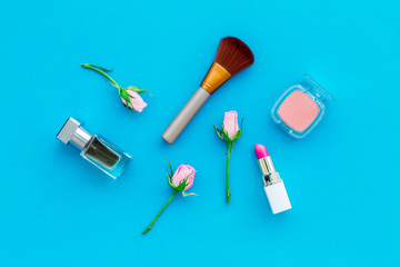 Rose tone cosmetics. Lipstick, bulk, eyeshadow, perfume among rose flowers on blue background top view