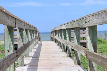 Fototapeta premium Perspective photograph of wood boardwalk railing beach access horizon blue sky, turquoise ocean and green grass.
