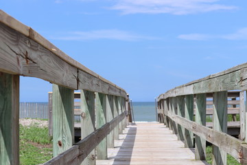 Fototapeta na wymiar Perspective photograph of wood boardwalk railing beach access horizon blue sky, turquoise ocean and green grass.
