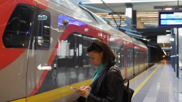 Underground modern railway station, a platform with girl looking at mobile, touching touch screen and yellow orange empty regional train on path, black tunnel stretching away, side view, stock video.