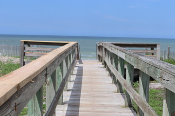 Fototapeta premium Perspective photograph of wood boardwalk railing beach access horizon blue sky, turquoise ocean and green grass.
