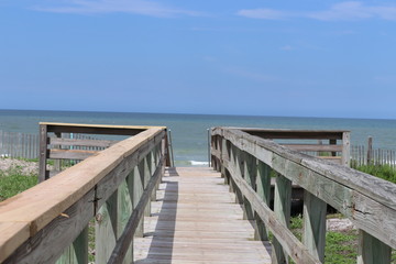 Perspective photograph of wood boardwalk railing beach access horizon blue sky, turquoise ocean and green grass.