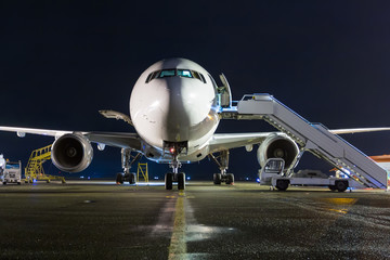 Front view of white wide-body passenger airplane with a boarding steps at the night airport apron