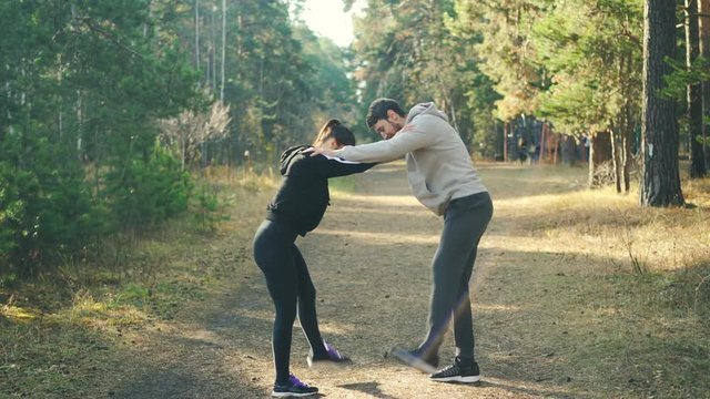 Man and woman are enjoying sports in park holding each other's shoulders and moving legs warming-up joints standing on path with green trees around them.