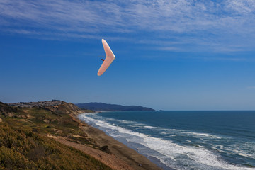 Hang gliding man flying above ground and ocean on a white wing at Fort Funston in San Francisco,...