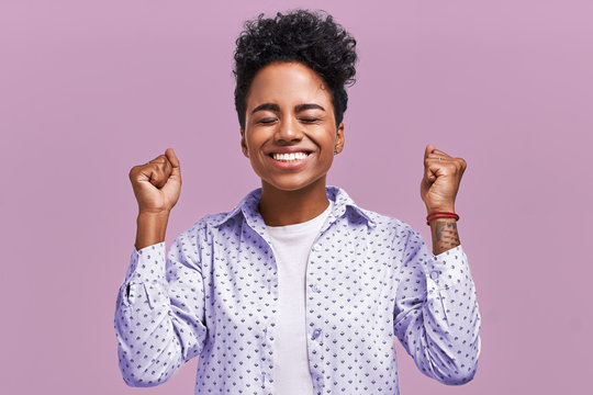 Horizontal shot of happy mixed race female clench fists with happiness, being satisfied with result of game, shout for their favorite team, have joyful expressions, isolated on lavender background