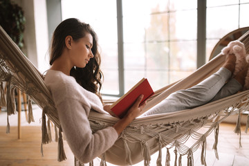 Dark-haired girl dressed in pants, sweater and warm slippers reads a book lying in a hammock in a...