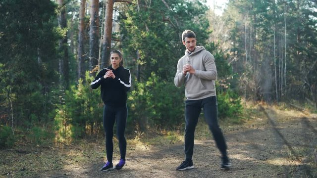 Beautiful couple young woman and her handsome boyfriend are doing physical exercises outdoors in park together doing squats and smiling. People and health concept.