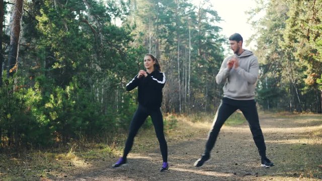Cheerful students are doing sports together in park squatting and smiling enjoying exercises and nature. Man and woman are wearing modern tracksuits.