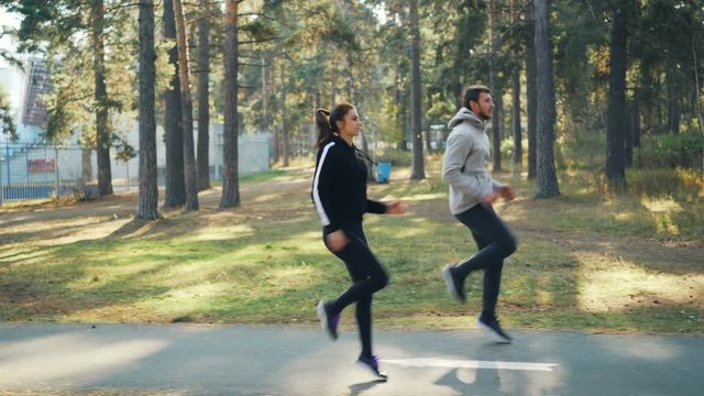 Side view of good-looking young people girl and guy doing sports in park running and jumping together on road on warm sunny autumn day. Nature and activity concept.