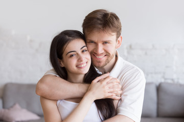 Fototapeta na wymiar Head shot portrait of loving embracing happy couple in living room, smiling boyfriend standing behind attractive girlfriend, hugging, man and woman holding hands, family portrait, look at camera