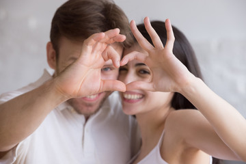 Naklejka premium Head shot portrait of loving happy couple showing heart, smiling boyfriend and attractive girlfriend looking through heart shape made with their fingers, funny family portrait, look at camera