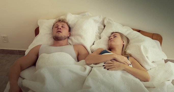 A young couple asleep in bed as the woman wakes up and is visibly annoyed by her partner's loud snoring, she covers her ears with her pillow