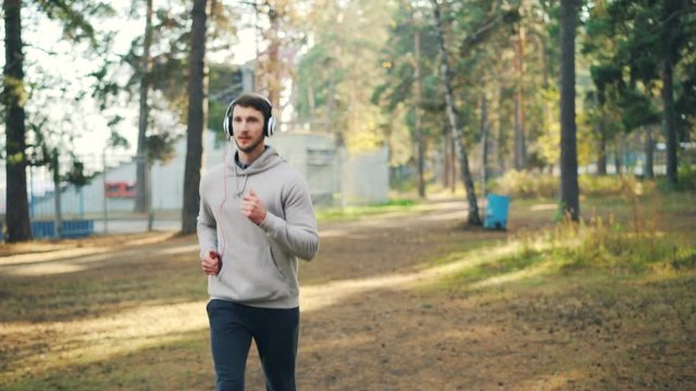Portrait of student jogging in park, listening to music with headphones and sometimes looking at camera. Guy is wearing trendy sportswear and using modern device.