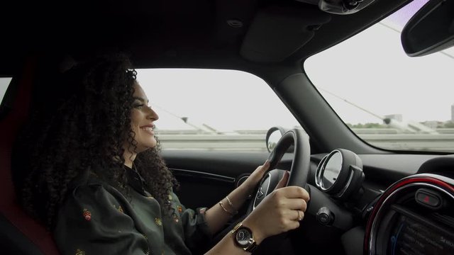Busy woman with curly hair driving a mini cooper and talking over speaker phone with joy
(medium shot)