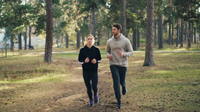 Dolly shot of attractive girl and guy jogging in park together focused on practice. Woman is wearing sweatshirt and leggings, man is wearing tracksuit.