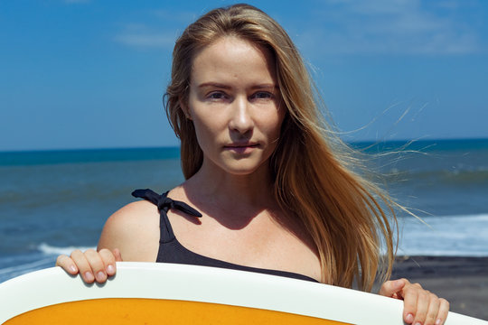 Surfer girl in bikini with surfboard posing on black sand beach. Face portrait. Young woman look at sea surf and waves. Active people in sport adventure camp, activity on summer family vacation