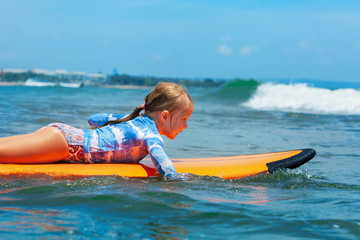 Happy baby girl - young surfer paddle on surfboard with fun on sea waves. Active family lifestyle, kids outdoor water sport lessons and swimming activity in surf camp. Beach summer vacation with child