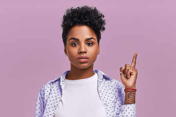 Shocked mixed race dark skinned female indicates upwards with puzzled expression, demonstrates something strange, wears fashionable clothing, on lavender studio background. Advertisement concept