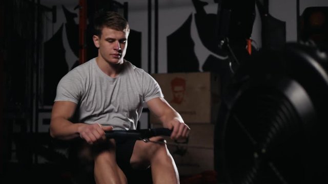 Young athlete doing rowing exercises in dark concept gym. Man boxing crossfit training machine power professional bodybuilder