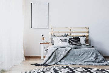 Mockup of empty poster in white bedroom interior with grey wooden bed with cushions. Real photo