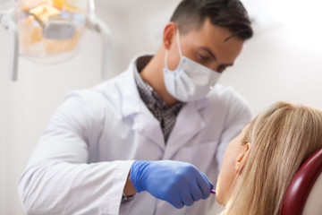 Fototapeta premium Professional dentist working at his office, examining teeth of a female patient. Male dentist in lab coat and medical mask checking teeth of a woman, copy space. Professionalism, experience concept