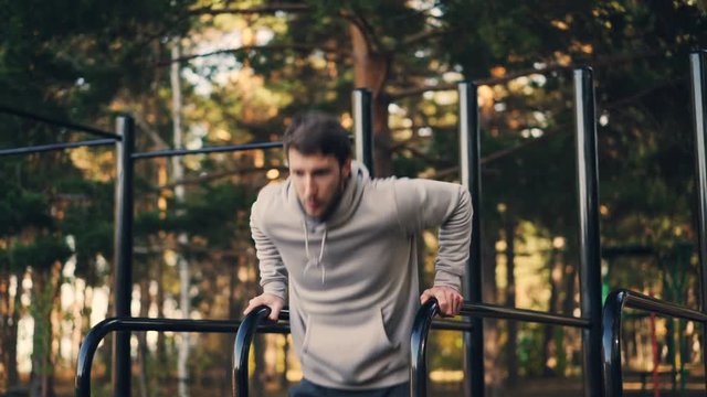 Strong young man is doing triceps dips on parallel bars in city park exercising outdoors in the morning in summer. Active lifestyle, workout and nature concept.