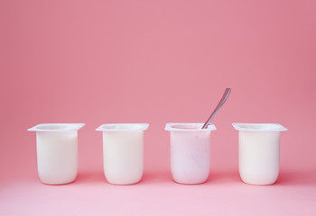 Four yogurts in white plastic cups on pink background with copy space. Strawberry pink yoghurt with spoon in it. Minimal style. Concept of better choice.