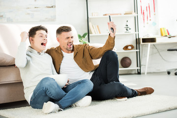 excited father and teen son watching match on tv and holding bowl of popcorn at home