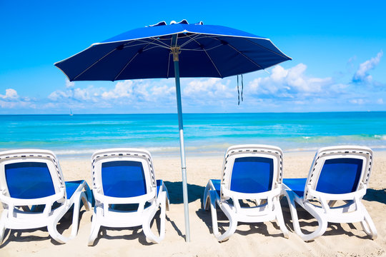 Sun loungers near blue umbrella on the sandy beach by the sea and sky. Vacation background. Idyllic beach landscape.