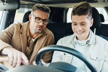 handsome father pointing on something while teaching smiling teen son driving car