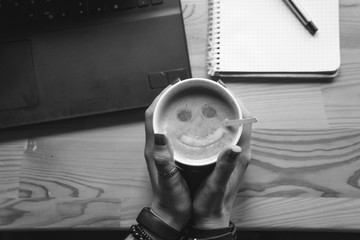 Top view. Student woman holds a hot cup of tea with sea buckthorn Hippophae and a slice of orange. happy smile in a cup. Workplace, notebook with pen and laptop, the working process, black and white