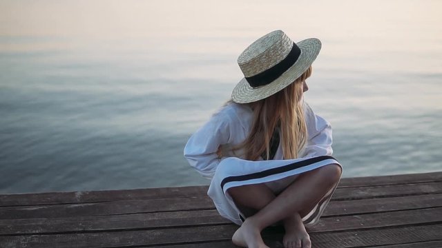 A little girl in a white dress sits on a wooden pier and looks at the sunset. Slow motion.