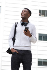 happy young african american man with bag holding on cellphone outside