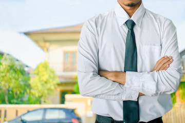 Man business salesman Sell house wearing necktie arm crossed On house and tree blurry background.For home business contracting or agreement and home warranty image.