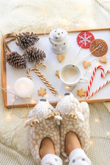 Fototapeta na wymiar Overhead view of girl feet in soft cozy deer slippers and tray with cappuccino, candle, lollipop with ginger man, Christmas candy cane, star cookies, decorative light, pine cones with garland on fur r